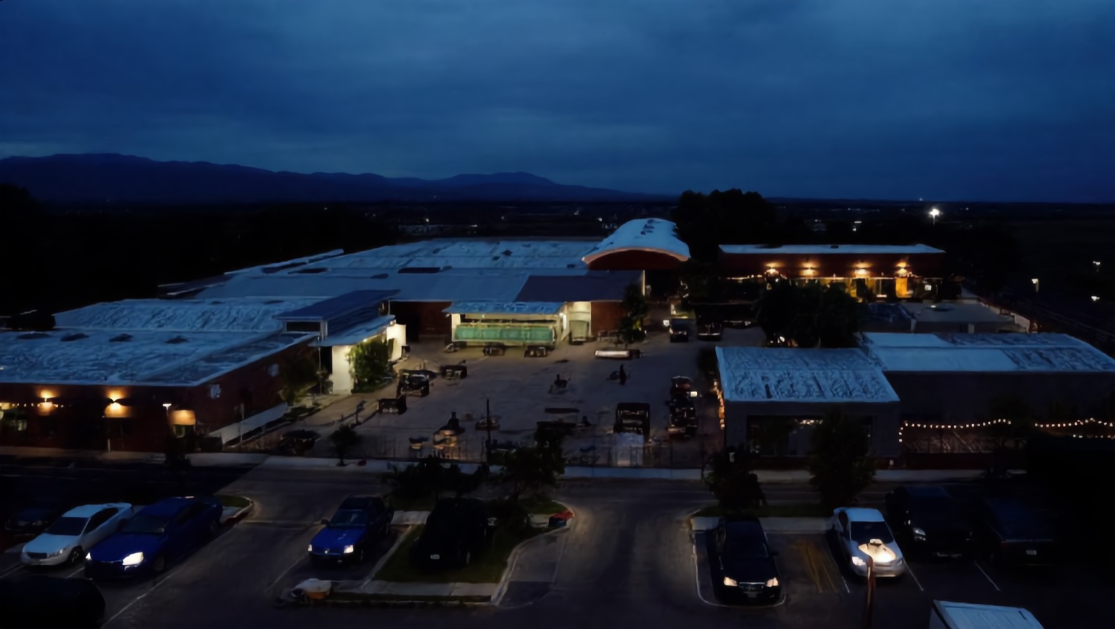 Concept render — the real five-building warehouse campus at dusk, matched to the property survey: low arched roofs, the lit central yard, mountains beyond